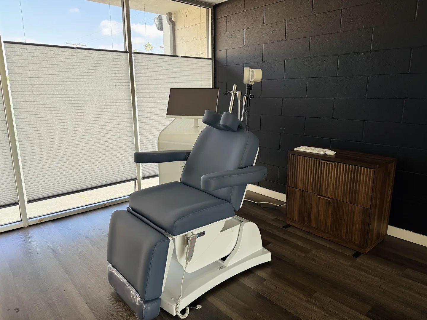 The TMS treatment chair in the NeuPath Long Beach clinic: a navy reclining chair with a headrest and monitor, beside a window with privacy shades